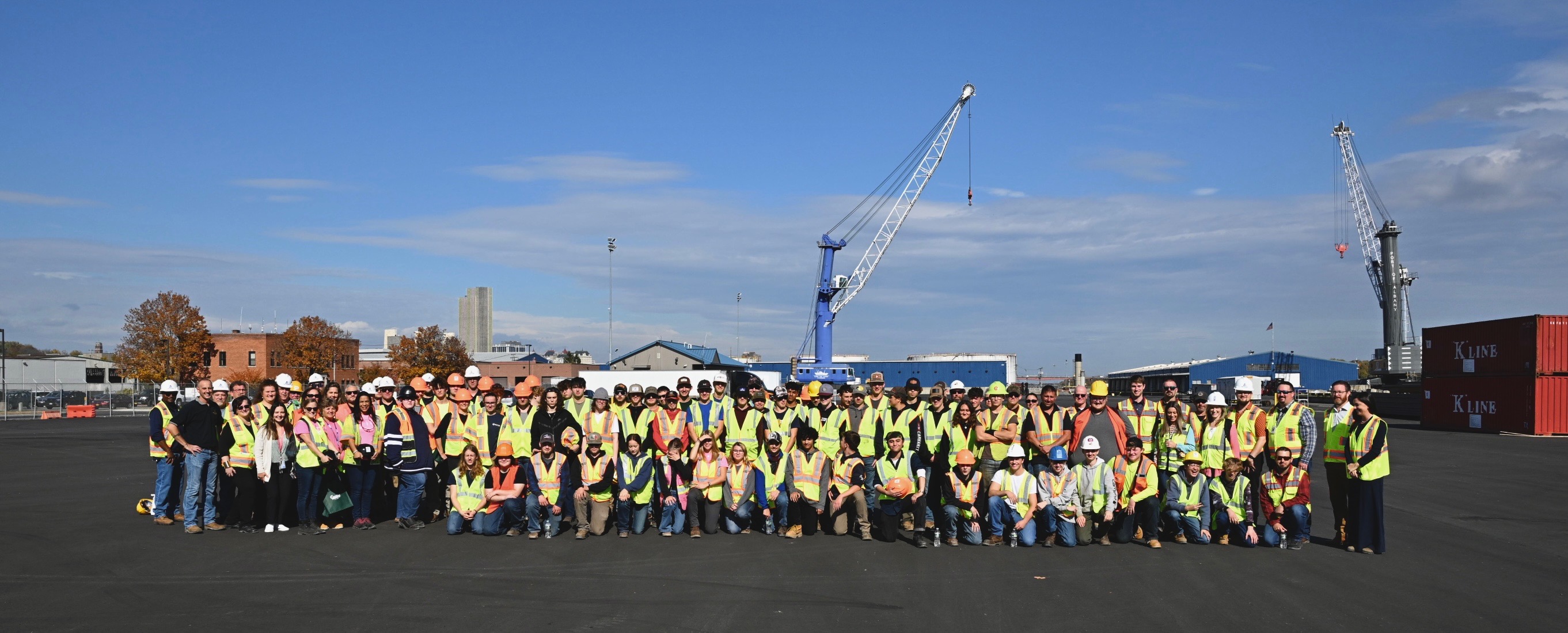 A large group of students, all in reflective gear, pose for a photo under cranes.