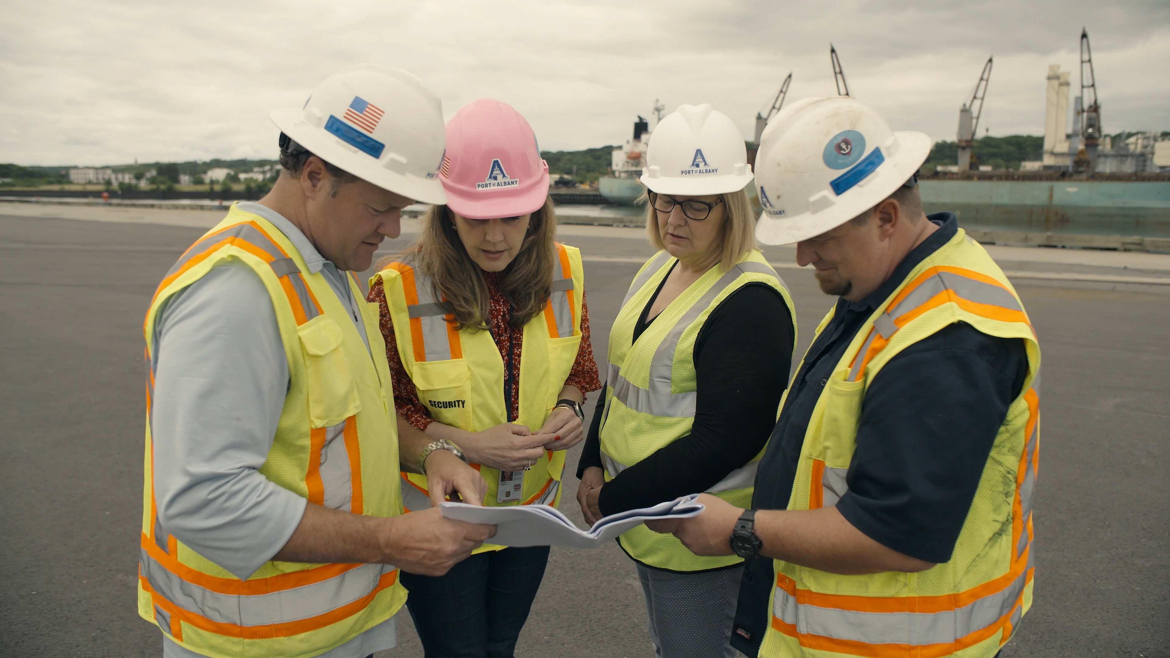 Four people wearing hardhats and safety vests look over a document together.