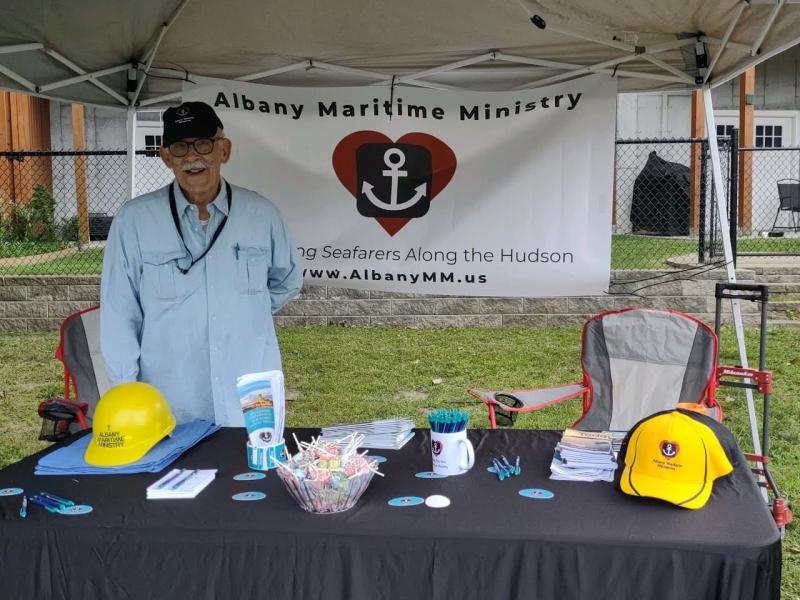 A man stands behind a booth with the Albany Maritime Ministry banner behind him, there are handouts and treats on the table in front of him.