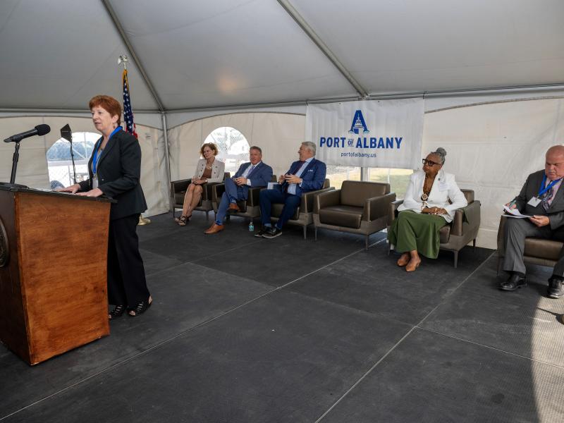 Albany Mayor Kathy Sheehan stands behind a podium addressing a crowd, the Port of Albany logo hanging on a banner behind her.