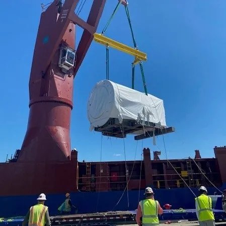 3 stevedores holding secure lines to large generator being lifted aboard a ship by crane