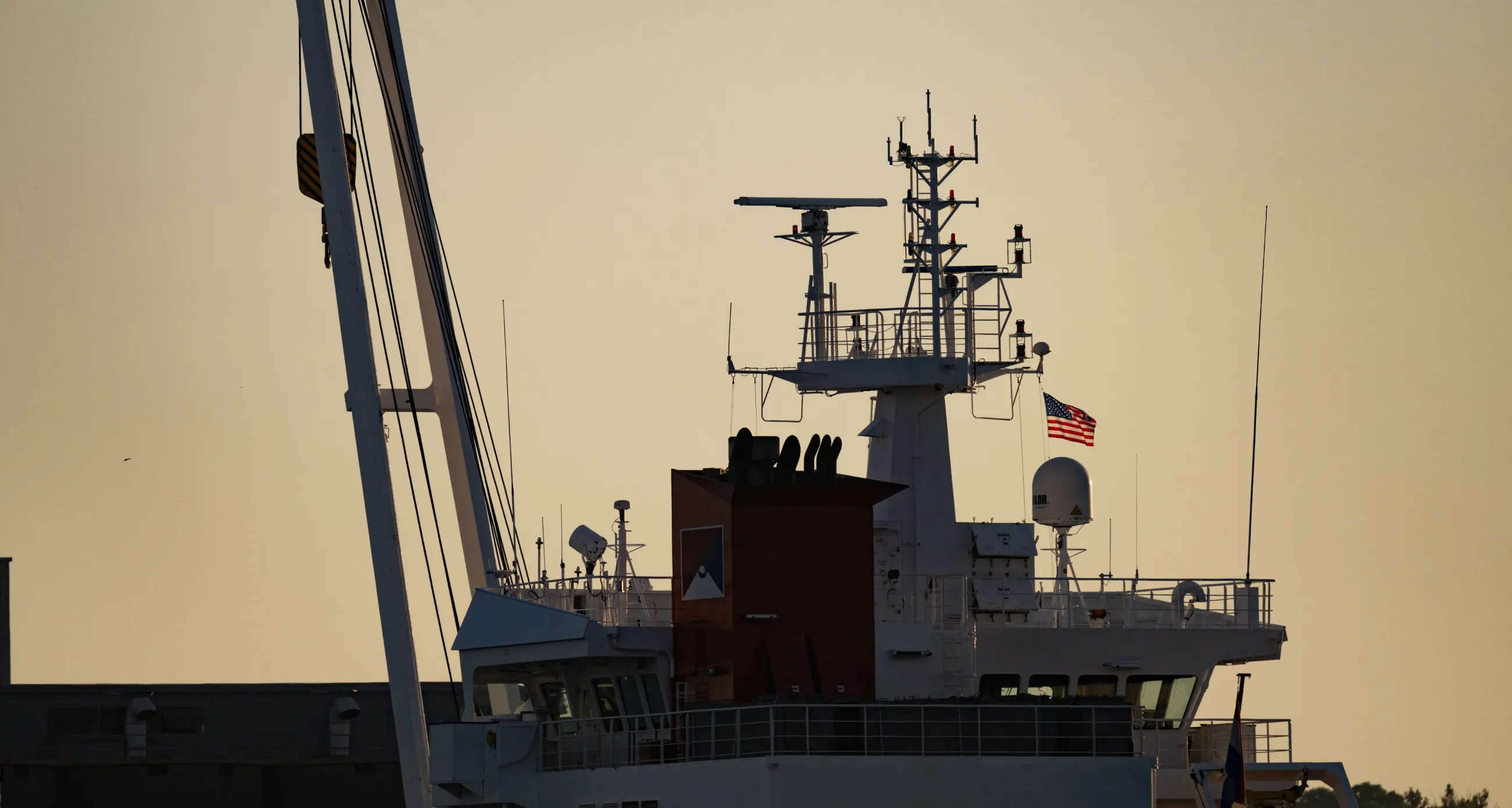 Ship sailing away in the sunset, an American flag can be seen backlit by the sun, flying over the ship. 